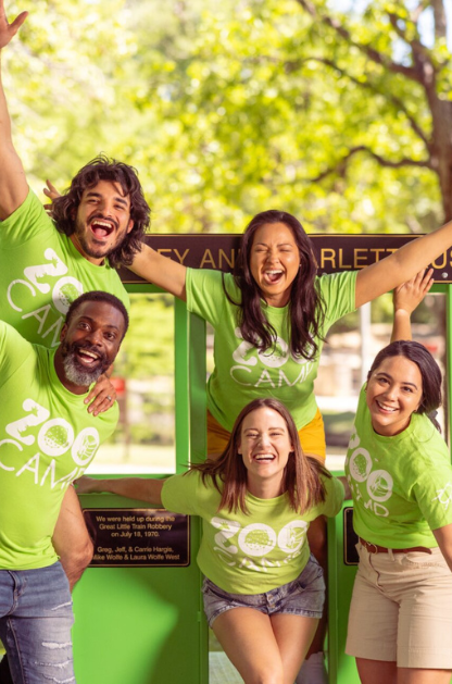 San Antonio Zoo camp counselors pose smiling in green camp shirts with arms raised, celebrating outdoors